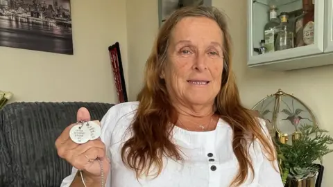 Angie Stone, a women in her 60s, with long brown hair, is holding two identification disks, on a chain, stamped with Gregg Stone's Army number, name and religion.