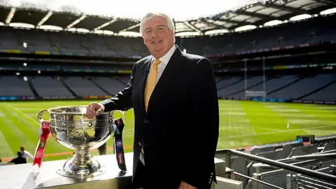 Getty Images A man with short grey hair stands with his right arm resting on a large silver bowl trophy, with red and navy ribbons around the trophy's handles. Behind him is a large empty stadium with a large green pitch in the centre. The man is wearing a black suit, white shirt and yellow tie. 