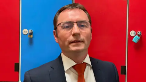 A headteacher - a middle-aged man a dark coloured suit jacket with a white shirt and red tie - stands in front of blue and red lockers. He is wearing thin-framed glasses.