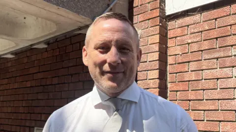A man with stubble, wearing a white shirt, standing in front of a red brick wall.