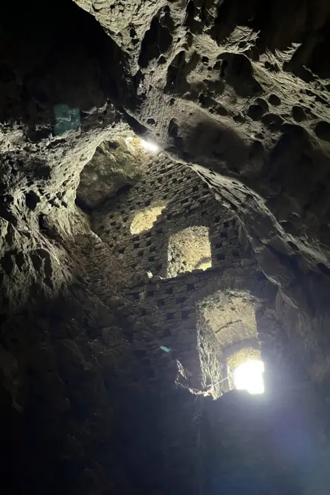 Nicole Barley Interior view of a cave with a curved, textured sides with small holes. A vertical brick-wall inside, with narrow openings letting in bright daylight that contrasts sharply against the dark, enclosed space.