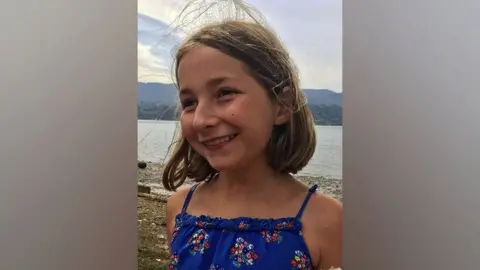 A 13-year-old girl, Martha Mills, is wearing a  blue sundress and looking happy. There is a lake and mountains in the background.