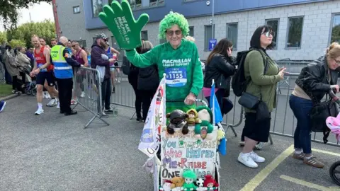 A man dressed in a green Macmillan Cancer Support jumper wearing a curly green wig and with a large green foam hand on his left arm is in the centre of the image. In front of him is a pram with various flags and toys. He is in front of a barricade which marks a race site, and there are race supporters and runners in the background.