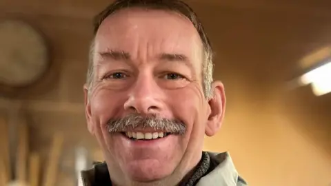 A man with short dark and grey hair and a grey moustache smiles at the camera with a wooden wall background behind him with a clock and what appears to be tools hanging on the wall. 