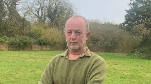 Supplied Johnnie Shand Kydd standing in a large green field which is surrounded by trees. He is wearing a green jumper and looking directly into the camera.