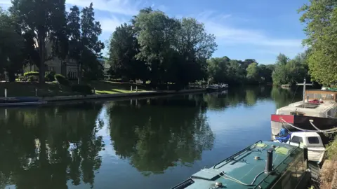 Vas Charitos  TUESADY - Boulter’s Lock at Maidenhead looking gorgeous in the sun. There are two boats moored on the right bank, a narrowboat with a green roof and a larger white narrow boat. There are trees overhanging the water and there are reflections of them in the water. On the opposite bank stands a two-storey house surrounded by trees and green grass.