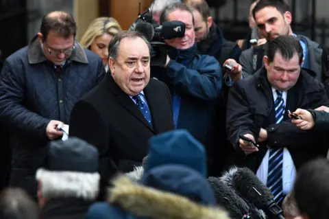 Getty Images A man in a dark suit and a blue tie, surrounded by reporters and cameramen