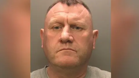 Paul Irwin looking directly into the camera in a police custody photograph. He has short dark hair and is wearing a grey round-necked top.