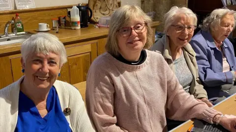 Four women sat next to each other smiling. The woman on the far left has a bright blue top and blue earrings, next to her is a woman wearing a pink knitted jumper and glasses. Next to that woman is a women wearing a blue top and beige fleece with glasses. On the far right is a woman wearing a purple jacket. They are sat inside with a sink behind them and milk, coffee and a kettle. 