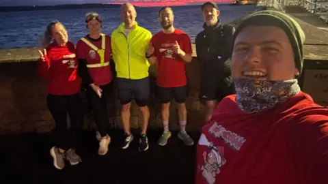 Ryan O'Shea A group of runners pose for a selfie at night as the sun sets creating a red skyline over the sea behind them. They are smiling and one runner is giving a thumbs up sign to the camera and another is giving a peace sign.