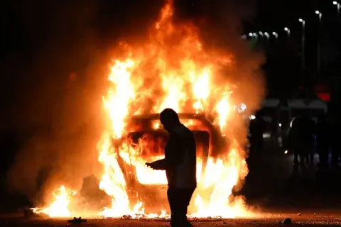 A silhouetted figure of a man looking at his mobile phone stands in front of a burning car which is on its roof 
