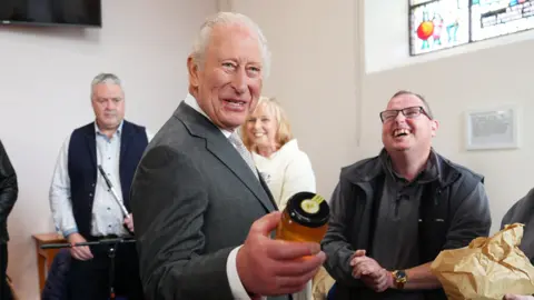 The King wears a dark-grey suit, white shirt and grey tie. He holds a clear jar of honey in his left hand. Her laughs as three people stand behind him. They are inside a building with white walls. A TV is over the Kings right shoulder and a stain-glass window is over his left.