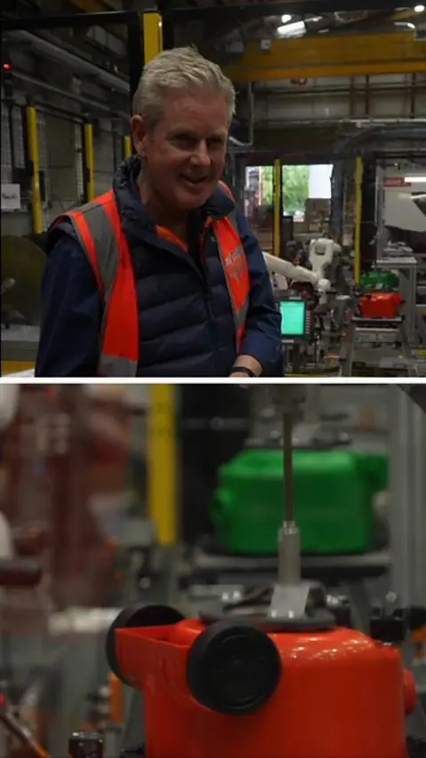A man stands in a factory which manufactures vacuum cleaners. He is wearing an orange high vis vest on top of a navy blue puffer jacket.