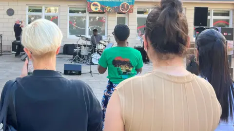 Outdoor community event outside a cream-colored building with a balcony, where a small band performs on a ground-level stage while a crowd watches from the foreground; speakers, instruments, and a banner hang behind the performers, and people sit at tables on the balcony above.