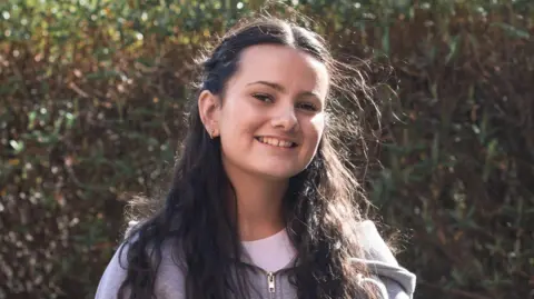A young woman with long dark hair which is pulled away from her face is smiling at the camera. She is wearing a grey hoodie top, with a white t-shirt underneath.