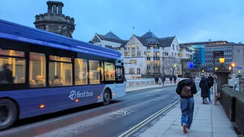 A purple First Bus travels across the Victorian iron Lendal Bridge while pedestrians walk along the pavement past the medieval Lendal Tower under an overcast winter sky.
