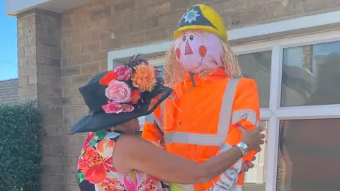 SUE UNWIN A lady wearing a floral dress and a wide-brimmed, black hat, decorated with flowers, holds a scarecrow that is wearing a hi-vis orange jacket and yellow helmet and is dressed to look like a fireman.