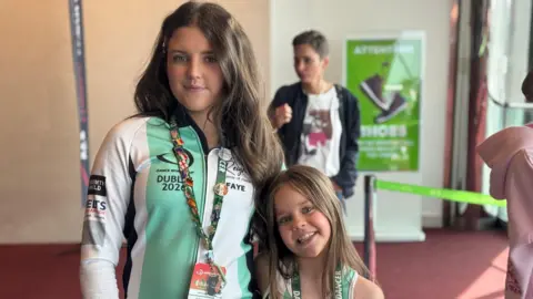 BBC Pictured are two girls at the Dance World Cup. On the left is team captain Faye and on the right is team member Mya. They are standing in a carpeted room with natural light coming in from the right. Faye is brunette, wearing a green and white striped DWC jacket and Mya is wearing a green vest top. They both have lanyards on with pins representing other countries and have brunette hair.