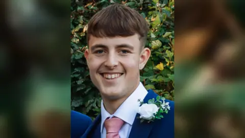 Ethan Powell wearing a navy suit and pink tie. He is smiling at the camera. 