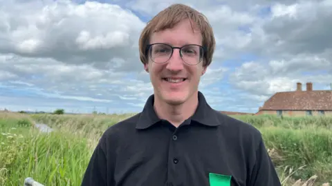 A man in a black shirt is standing by a river. He is wearing glasses and is smiling.