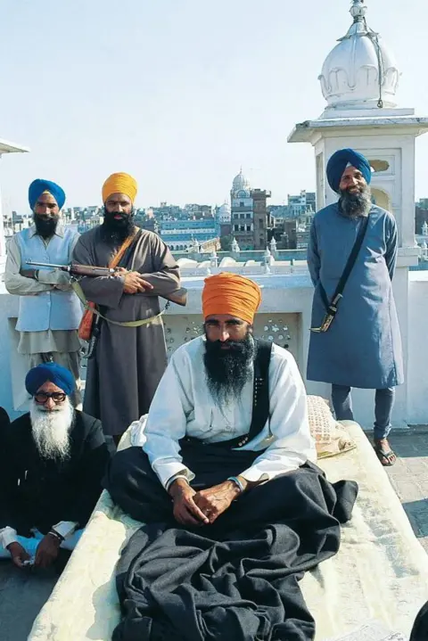 Raghu Rai/The India Today Group via Getty Images Sikh separatist leader Jarnail Singh Bhindranwale, wearing a turban ans surrounded by his armed supporters and guards, sitting on a cot.
