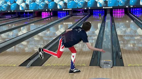 Stroud Sharks YBC A young boy wearing Union Jack patterned trousers and a navy blue bowling shirt. He is standing in a bowling alley and has just thrown a ball, which is rolling towards the ten pins at the end of the shiny wooden lane. Above the lanes are large electronic screens which display stacked blue bowling balls. The pins are illuminated by pink and blue LED lights.