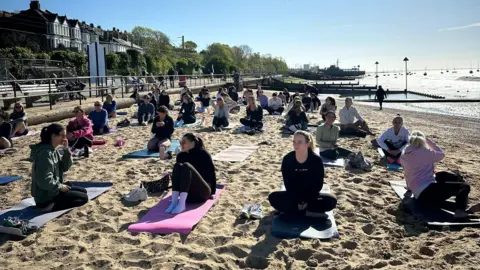 Alexandra Beard A dozens of people sitting on their yoga mats on a beach before or after a session, the sun is out