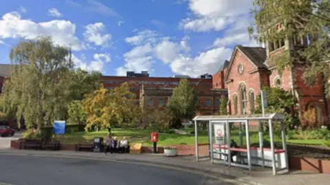 The outside of a red-coloured Victorian hospital building. In the foreground is a bus stop placed on the edge of the road, with pedestrians also going about their business.