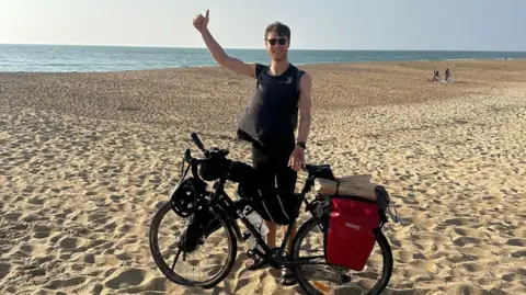 Ollie wears a black vest and shorts, and stands with his bike on a very wide sandy beach. The blue ocean can be seen behind him. He has one hand on his bike and the other raised in a thumbs up. 