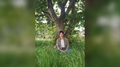Natalie Fée sat under a tree. She has her eyes closed and is surrounded by long grass.