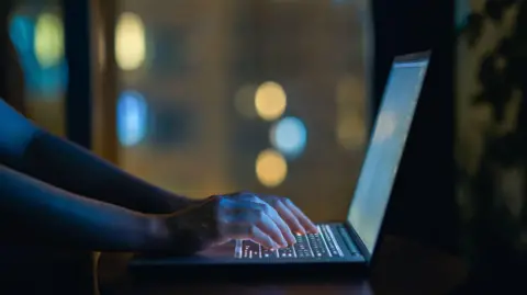 Getty Images A person is typing on a laptop. The laptop screen and keyboard is lit up. The room is dark. 