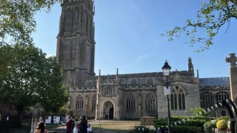 BBC St John's Church on a summer's day. Several people can be seen standing on the pavement besides the black railings outside. There are trees to the left and bunting to the right. 