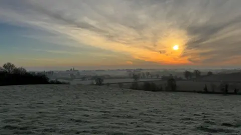 A patchwork of fields runs to the horizon. In the foreground, frost can be seen on the grass and in the distance mist covers the fields and trees. The rising sun creates a golden glow in the sky above the fields.