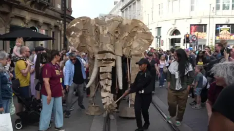A wooden elephant puppet moves through Manchester city centre with crowds around 