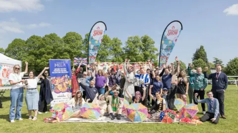 People gather at a previous Worcester Show, some are wearing costumes and some holding signs and banners that say Mela or Worcester Show. They have their arms in the air and there is a festival atmosphere, with a marquee behind, and they are looking happy. They're outside, on grass, and it is a sunny day.