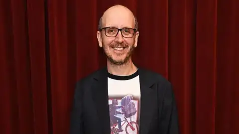 Dave Benett/Getty Images A midshot of Jack in front of a red velvet curtain, smiling to camera, wearing glasses and a black suit jacket with a white t-shirt bearing the iconic shot of the film ET with the boy riding a bicycle and ET in the basket