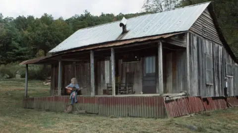 Getty Images Dolly Parton sits on the porch of a replica of the log cabin she grew up with