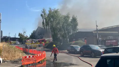 Shariqua Ahmed/BBC A firefighter is standing on a road in the middle of an industrial estate. Several cars are parked on the road. In the background, smoke is rising from a building.
