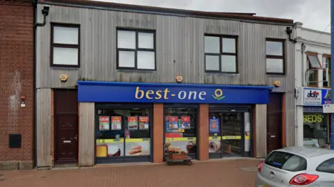 Google A shop with the words Best One on a blue background at the top of the ground floor outside. Part of a pavement and a silver car are visible in front of the store.
