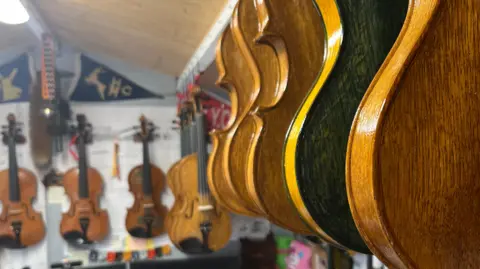 John Devine/BBC A number of handmade violins hang from a wall in a workshop with some tools visible around them. 