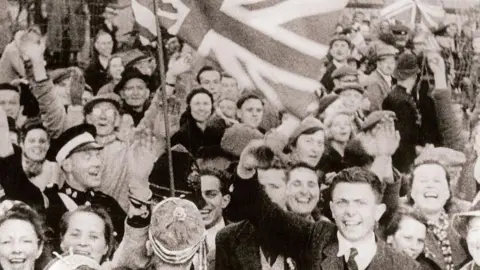 Black and white picture of people smiling as they celebrate the liberation of Guernsey on 9 May 1945. A large Union Flag is waving above the crowd.