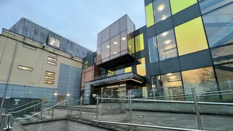 The exterior of Wiltshire County Hall, Trowbridge. The building has a modern facade with large square multicoloured panels in grey, orange and yellow. There are stairs leading up to the front of the building and the words county hall above the entrance