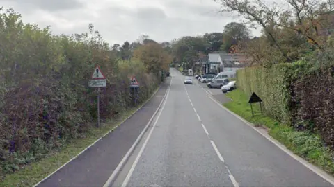 Google A view of the A595 road into Holmrook. On the left side of the road there is a 30mph sign and a warning sign for pedestrians crossing and a school. On the right side of the road, there are parked vehicles and the entrance to a garage.
