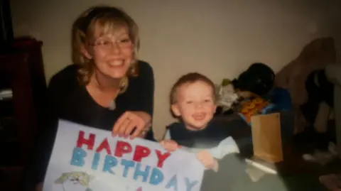 Alfie Watts A young Alfie Watts and his mother both smile as they both hold a big piece of paper bearing the words "happy birthday"