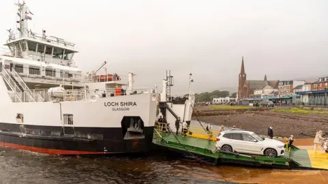 Getty Images A ferry docked at the coast, with a white car and seveal passengers disembarking from the boat. Various buildings and a church can be seen in the background.