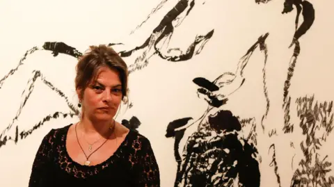 Reuters Tracey Emin is photographed in front of one of her artworks. Emin is wearing a black lacy top with two pendant necklaces and dangly earrings. Her hair is tied up and she is looking to one side. The painting behind her shows a woman in the nude and is painted all in black on an cream canvas. It is called "Distant Memory" and was shown in Vienna.