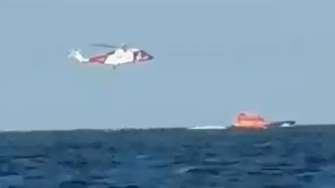 Diana Hare A helicopter hovers over an orange lifeboat on the water.
