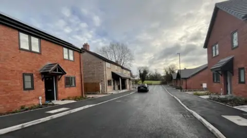 A newly paved road with brand new homes on either side. The homes are red brick detached houses apart from the back left one which is more of a brown/chalky colour. On the street is a solitary car and ahead of the houses is a field. There are a number of trees in the distance that have not got any leaves and a cloudy sky with patches of light blue.