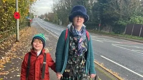 A woman wearing a flurry hat and a cardigan stands on a street holding hands with a young boy. In her other hand she is holding a dog's harness but her dog is not in frame. 