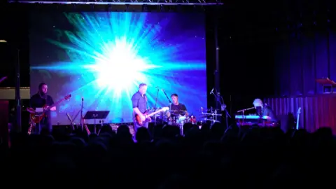 Four members of band playing instruments with a bright blue light behind on a screen. The image takes up most the screen. 